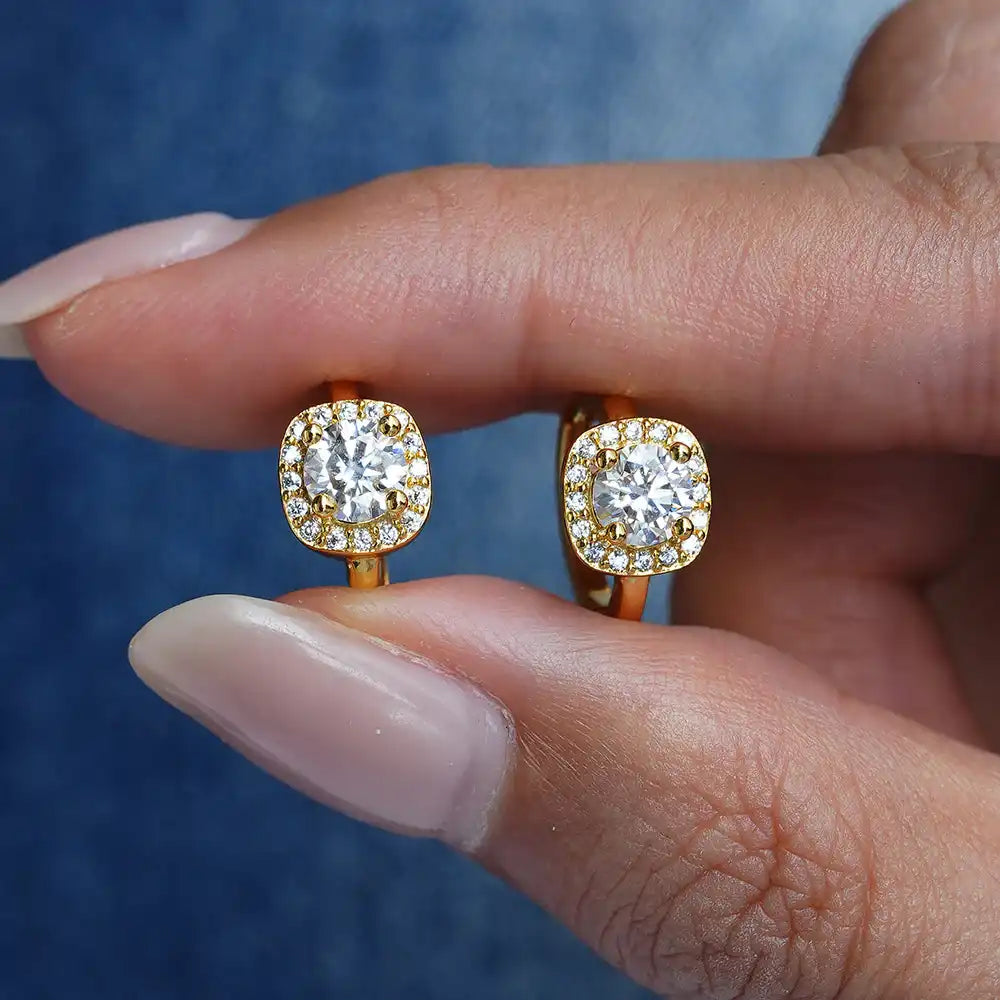 Gold moissanite halo earrings with round brilliant stones held by hand on blue background