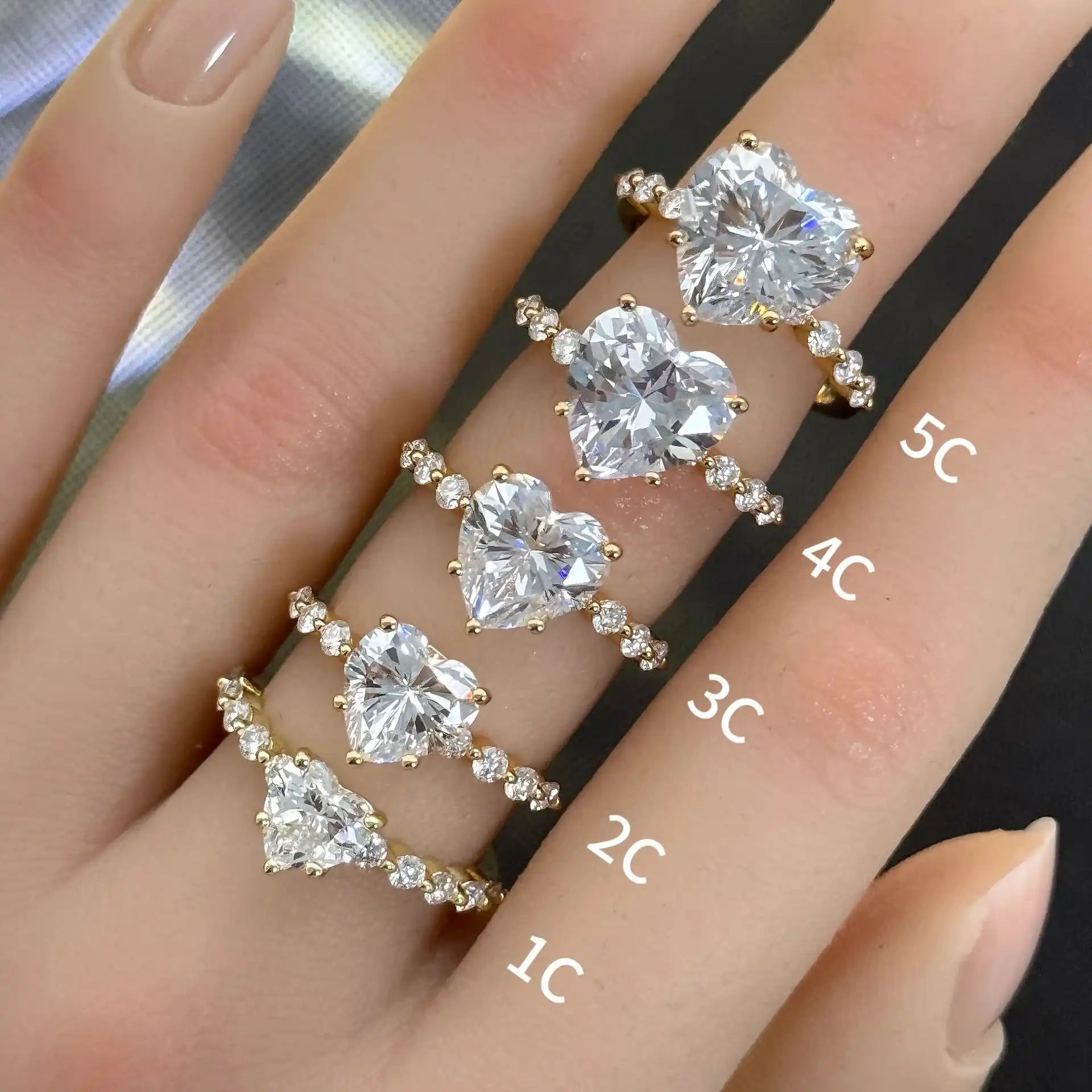 Hand wearing gold moissanite rings with heart-shaped stones in various carat sizes, labeled 1C to 5C.