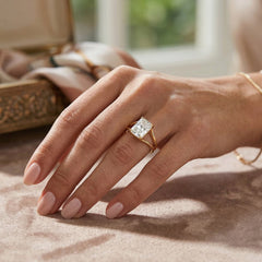 Hand wearing gold band moissanite ring with radiant-cut stone on soft beige surface