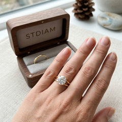 Hand wearing gold moissanite engagement ring with jewelry box and pine cone in background