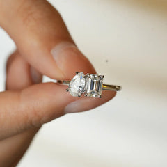 Hand holding a two stone moissanite ring with pear and emerald cut gems on a silver band.